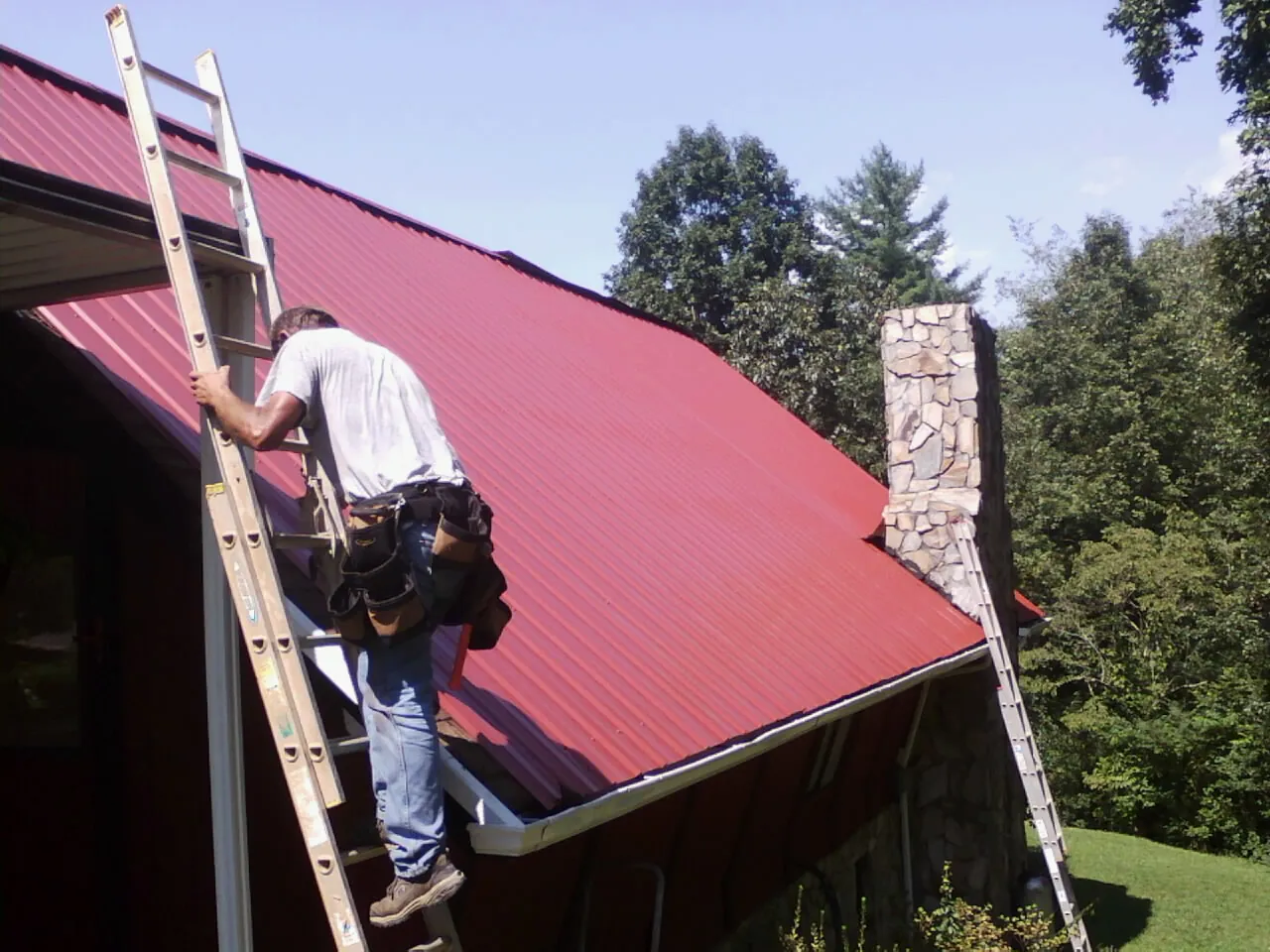 Roofer on a ladder beside a red metal roof and stone chimney — Soffit & Fascia Repair by Roofing Brothers in Tarawa Terrace